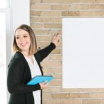 Emilie standing, holding a blue ipad and pointing at a blank piece of poster paper on the brick wall.