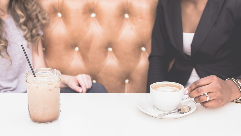 Two friends with coffees at a café banquette, mid-conversation — the unhurried meet-up where creative inspiration often sneaks in.
