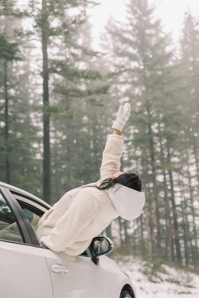 Person in a cream coat leaning out a car window with arm raised, snowy pine forest behind — long drives unlock creative inspiration.