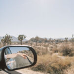 Side-mirror view of Joshua Tree desert, an arm reaching into the open air past cacti and boulders.
