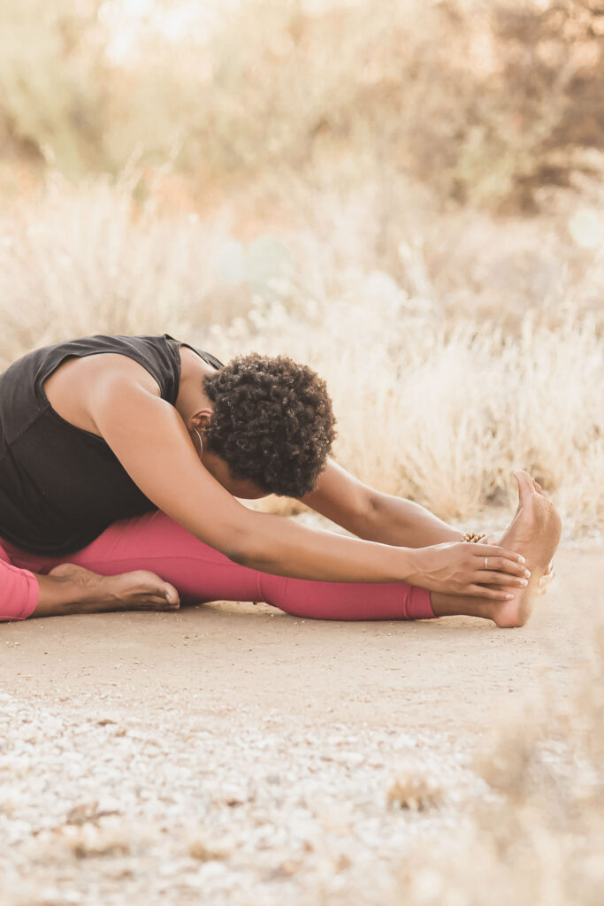 Woman stretching outdoors in a seated forward fold, reaching toward her foot, with dried golden grasses blurred in the background