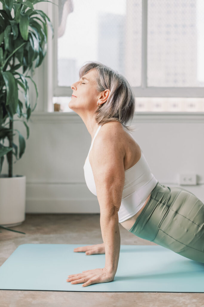 Older woman doing upward-facing dog yoga pose on a blue mat, eyes closed, near a bright window with a large leafy plant
