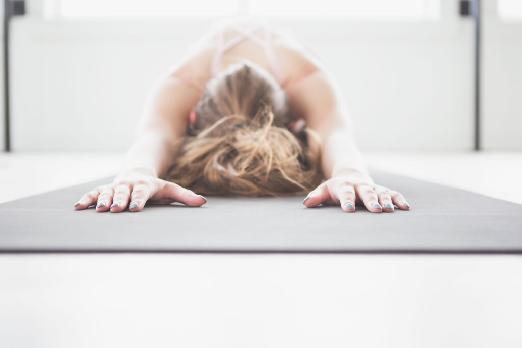 Woman in child's pose on a gray yoga mat, arms extended forward, hair down, photographed from the front in a bright minimal studio