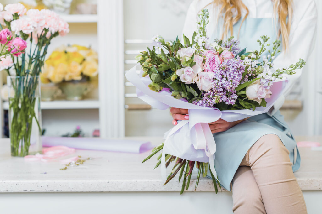 A florist in a light blue apron holds a lush bouquet of pink roses and lavender blooms inside a flower shop